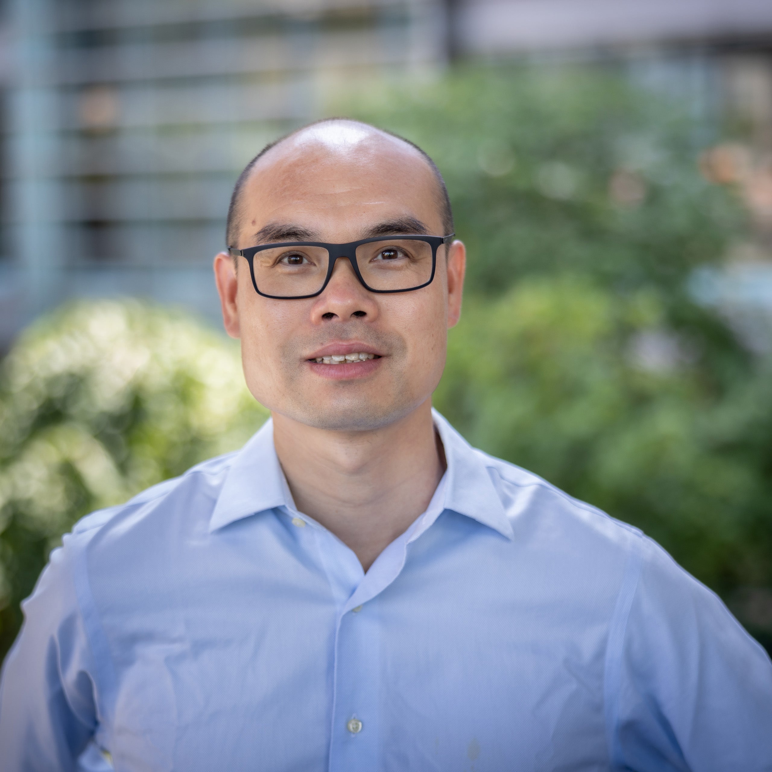 Guoshi Li's headshot. An Asian male with balding hair and brown eyes wearing glasses looking at the camera. He is wearing a light blue open-collard button-down shirt.