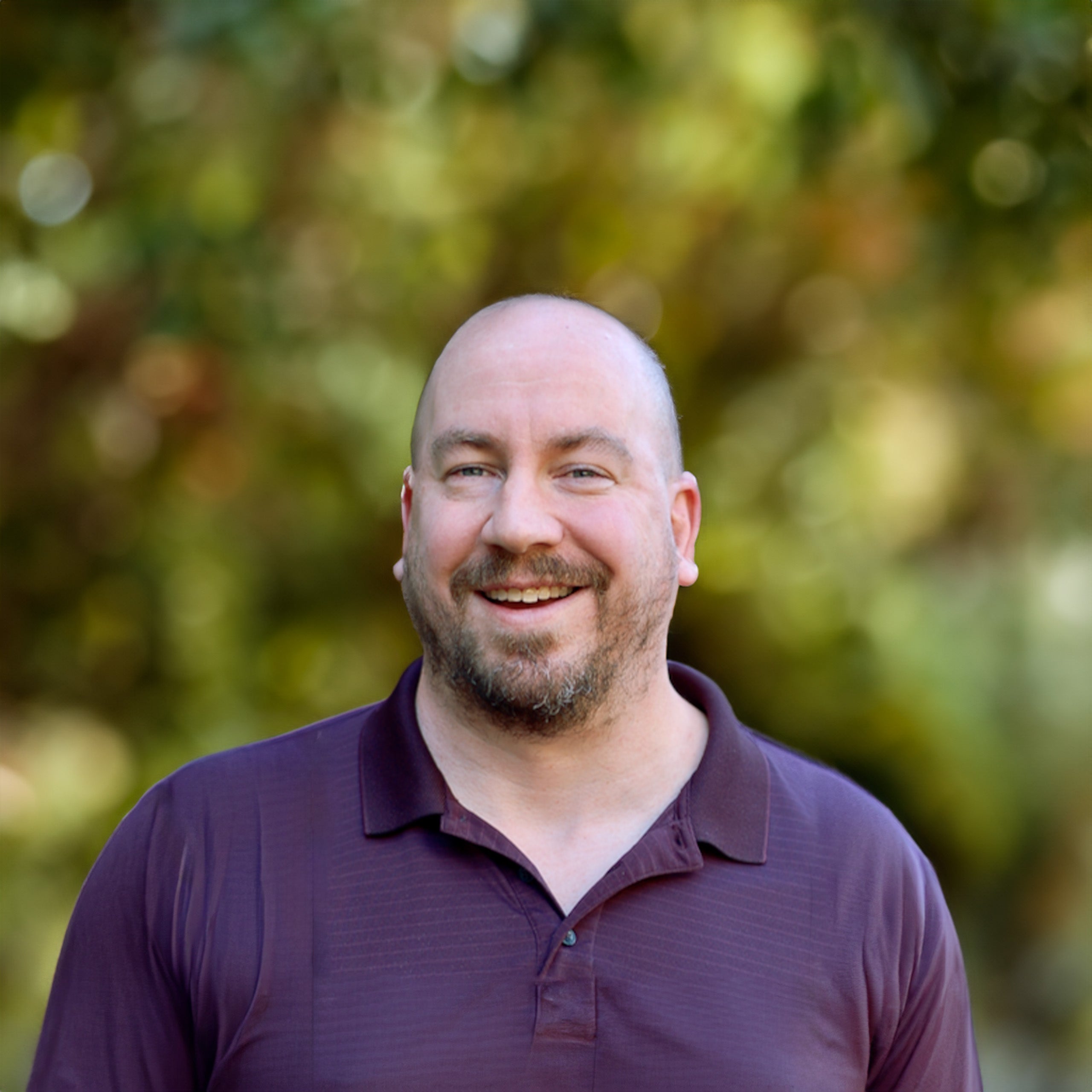 Thad Benefield headshot. Bald male with blue eyes and a light brown beard smiling at the camera. He is wearing a deep purple polo shirt.