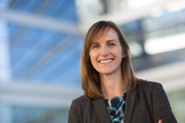 Louise Henderson headshot, white female standing in front of glass windows, shoulder-length brown hair, blue eyes, smiling at the camera in a dark gray jacket and blouse.