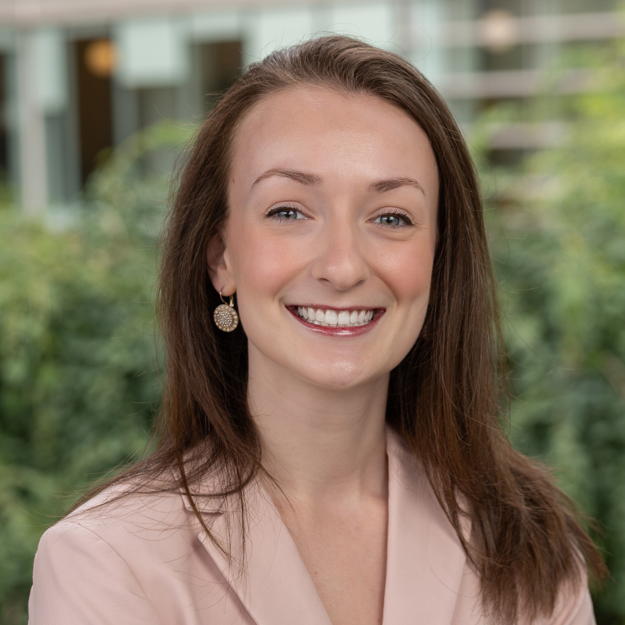 Headshot of Kristen Tignor, white woman with long, light brown hair smiling at camera. She is wearing a pink jacket and is standing against an outdoor backdrop.