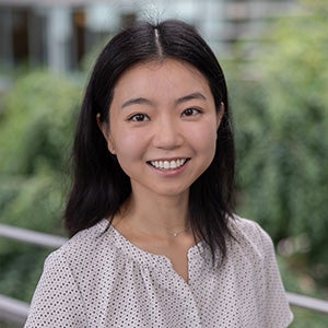 Dahyun Kang headshot. Asian female with long black hair, brown eyes, smiling at the camera. She is wearing a white blouse amd standing against an outdoor backdrop.