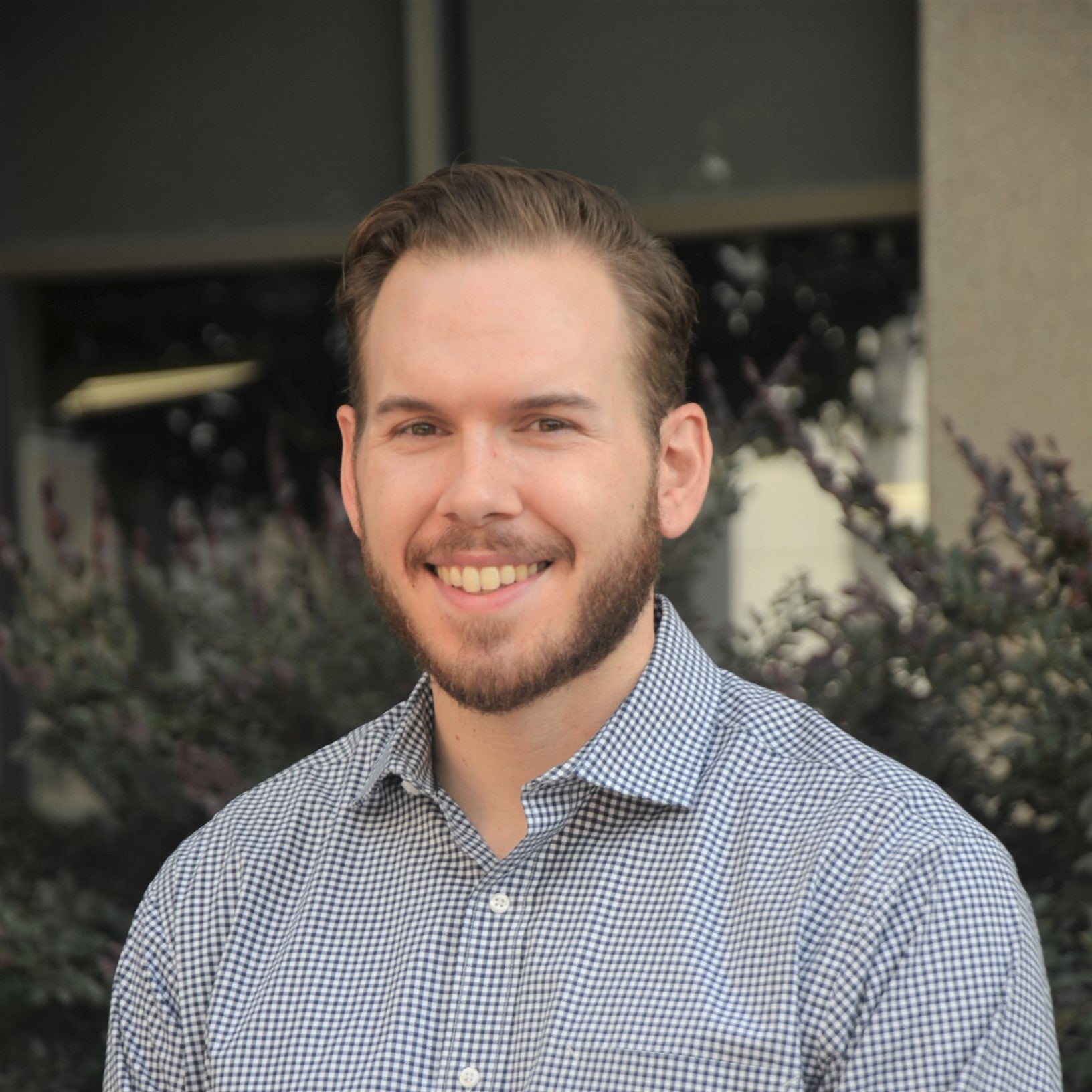 A male with short light brown hair, beard, and hazel eyes, smiling at the camera. He is wearing an open-collar, checkered button-down shirt. He is standing against an outdoor backdrop.