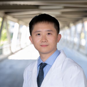 An Asian male with short black hair and brown eyes is looking at the camera. He is wearing a white doctor's coat, blue shirt and black tie. He is standing against an outdoor covered bridge.