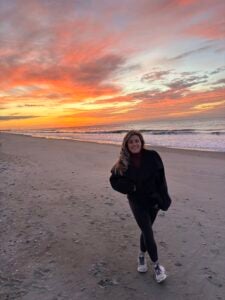 Young female with blond hair wearing black pants and sweater posing in front of a firy sunset ay the beach. 