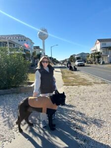White blond female smiling at the camera beside her black sherman Sheppard at the beach with a water tower beind her. 