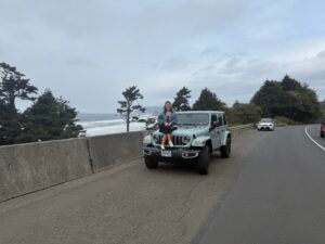 Female sitting on the hood of a blue jeep on the side of the road in a foreign country. 