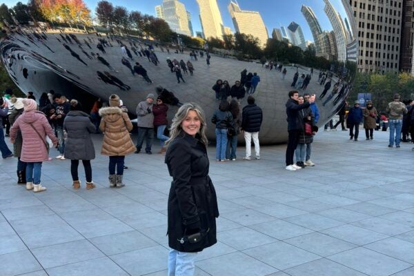 Female in jeans and a black coat smiling at the camera with the "Cloud Gate," a large, highly-polished, mirrored bean-shaped sculpture in Chicago.