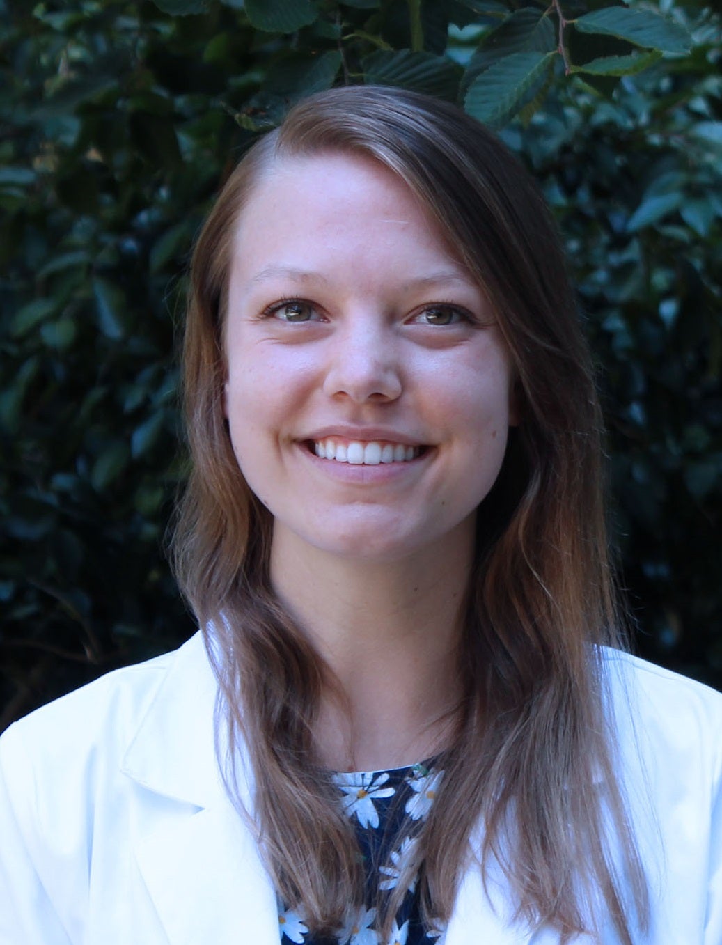 Professional head shot, outdoor background, white coat over office attire.