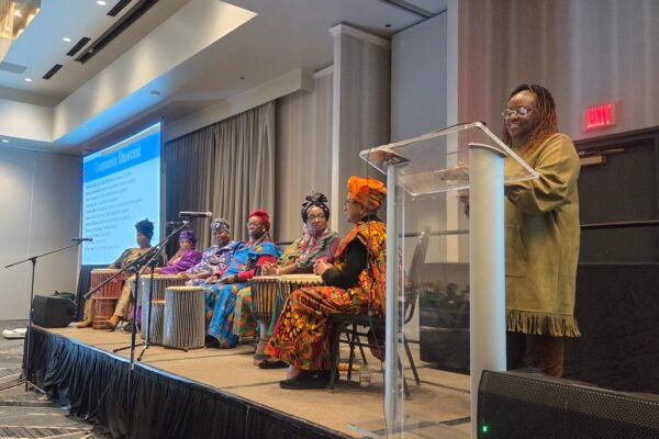 A photo of 5 women sitting on a stage with percussion instruments to the left of a woman smiling and standing at a podium in a conference room.