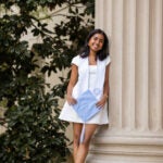 A student holding a blue graduating cap wearing a white dress leaning on a column outside posing for a picture