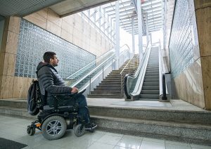 man in wheelchair in front of escalator and stairs with no accessible way upstairs.